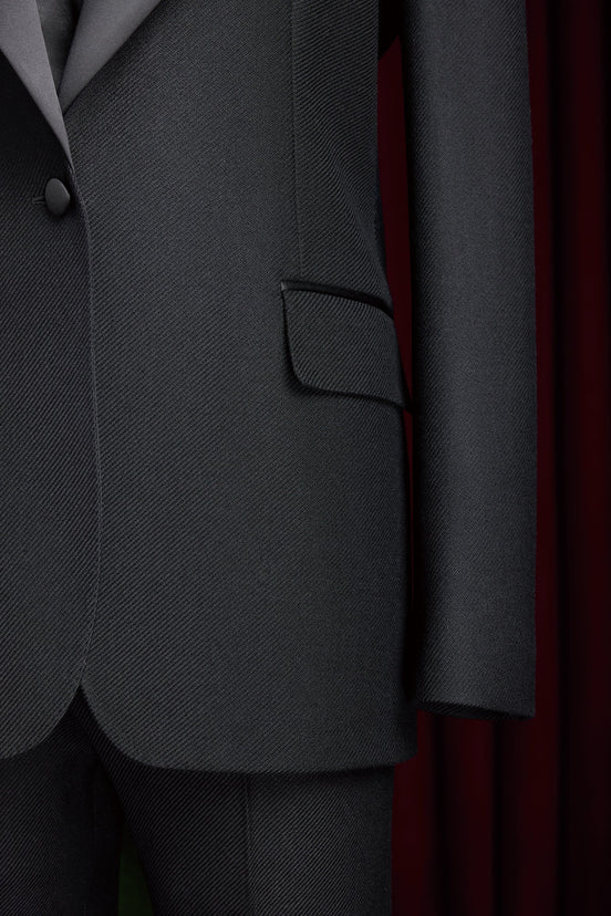 Close-up of women's black tuxedo suit with satin lapel, single-button closure, and flap pockets, showcasing luxurious tailoring and elegant design.