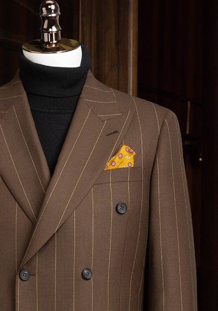 Close-up of a brown pinstripe men's suit jacket with double-breasted design, paired with a black turtleneck and accented by a vibrant orange pocket square for a refined, high-end look.