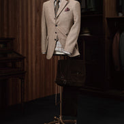 Elegant beige men's tweed blazer on a mannequin with a plaid shirt, floral tie, and pocket square, styled in a vintage-inspired tailoring shop.