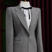 Women's tailored houndstooth suit on mannequin, featuring structured shoulders, fitted waist, and white blouse with black ribbon, exuding vintage elegance.