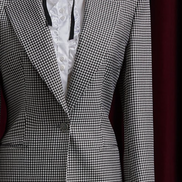 Women's tailored houndstooth suit on mannequin, featuring structured shoulders, fitted waist, and white blouse with black ribbon, exuding vintage elegance. Close up on peak lapels and button