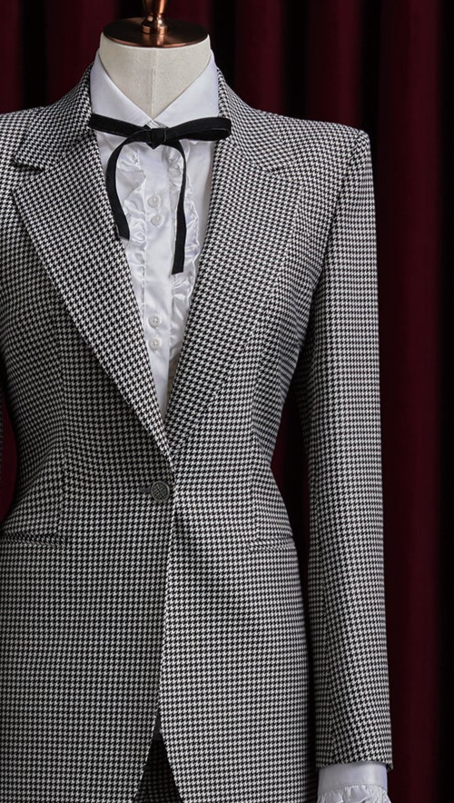Women's tailored houndstooth suit on mannequin, featuring structured shoulders, fitted waist, and white blouse with black ribbon, exuding vintage elegance. Close up on peak lapels and button